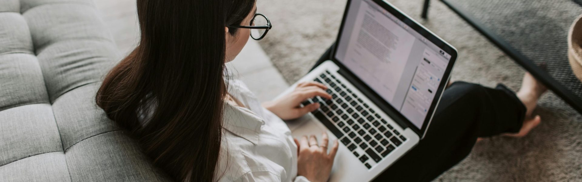 Woman working remotely with a laptop on the floor next to a sofa, enjoying comfortable home office setup.